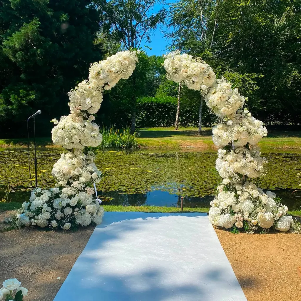 Wedding Arch White floral wedding arch over a light blue aisle runner by a pond in a garden setting.